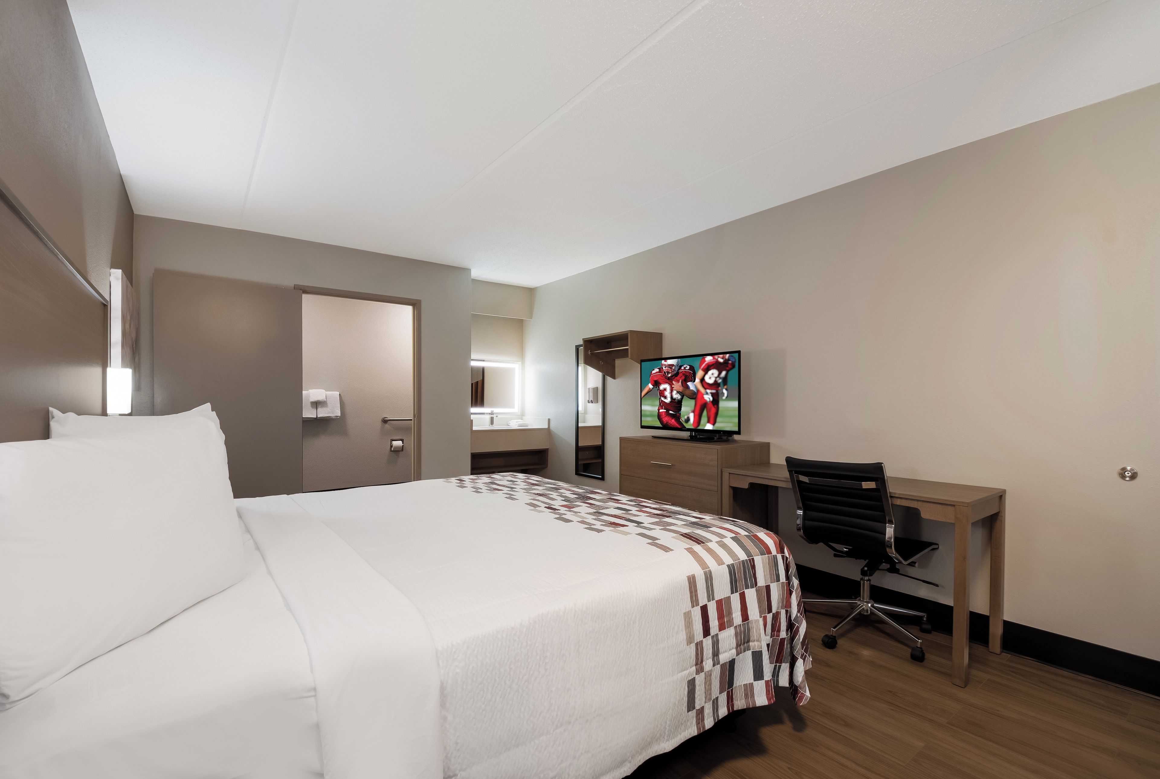 A hotel room with a bed, a desk, a chair, a television, and a bathroom visible through an open door.