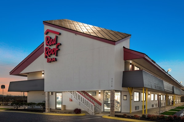 Ett Red Roof Inn-hotell med en modern design, präglat av en tydlig färgskala i rött och vitt, ett takförsett utomhusområde och en trappa som leder till entrén.
