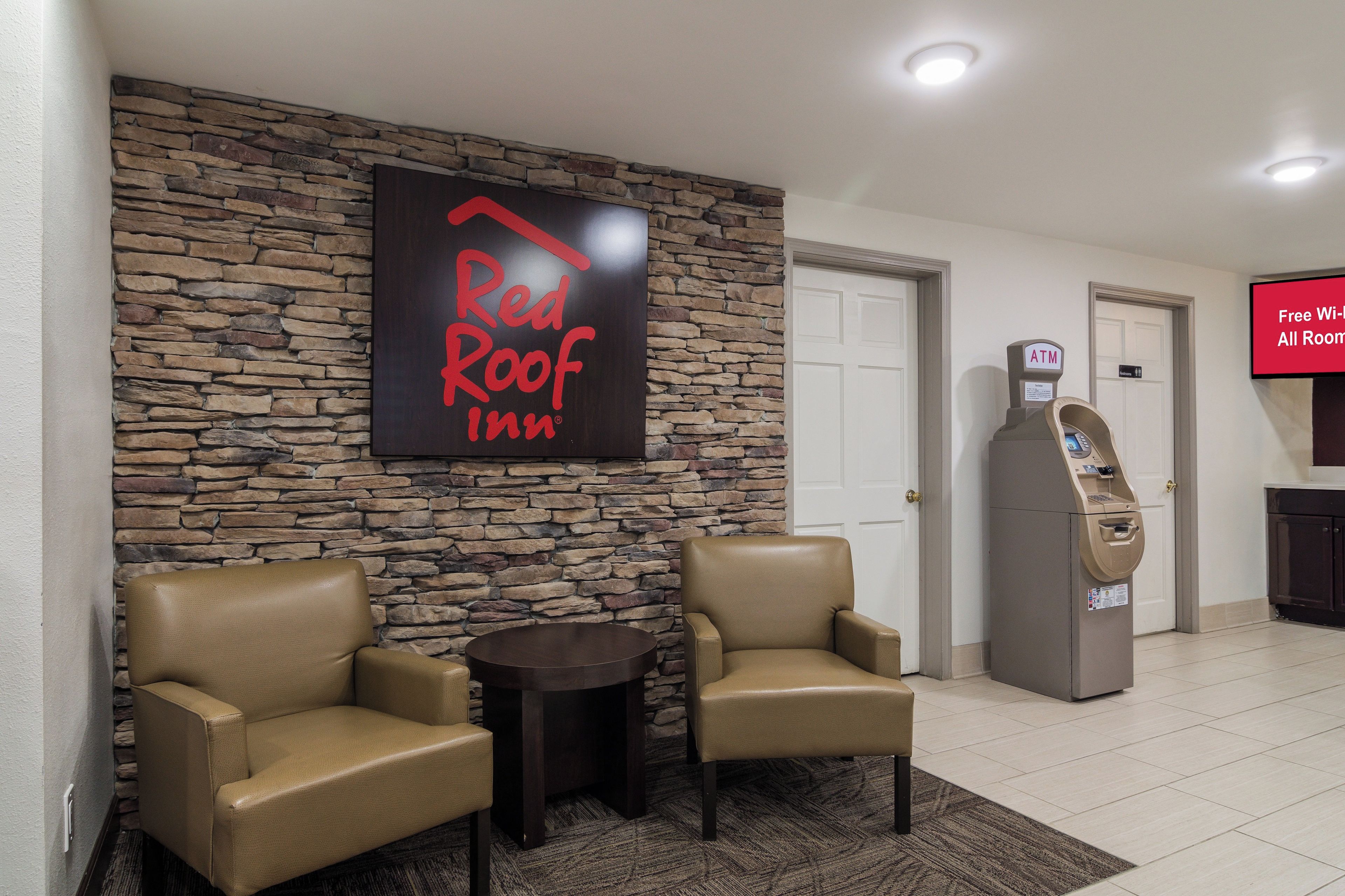 A Red Roof Inn lobby with stone walls, leather armchairs, a small table, an ATM, and a sign indicating free Wi-Fi in all rooms.
