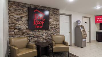 A Red Roof Inn lobby with stone walls, leather armchairs, a small table, an ATM, and a sign indicating free Wi-Fi in all rooms.