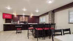 Uma área de refeições com uma cozinha ao fundo, uma TV de tela plana exibindo 'Wi-Fi Gratuito em Todos os Quartos' e uma parede de destaque em pedra.