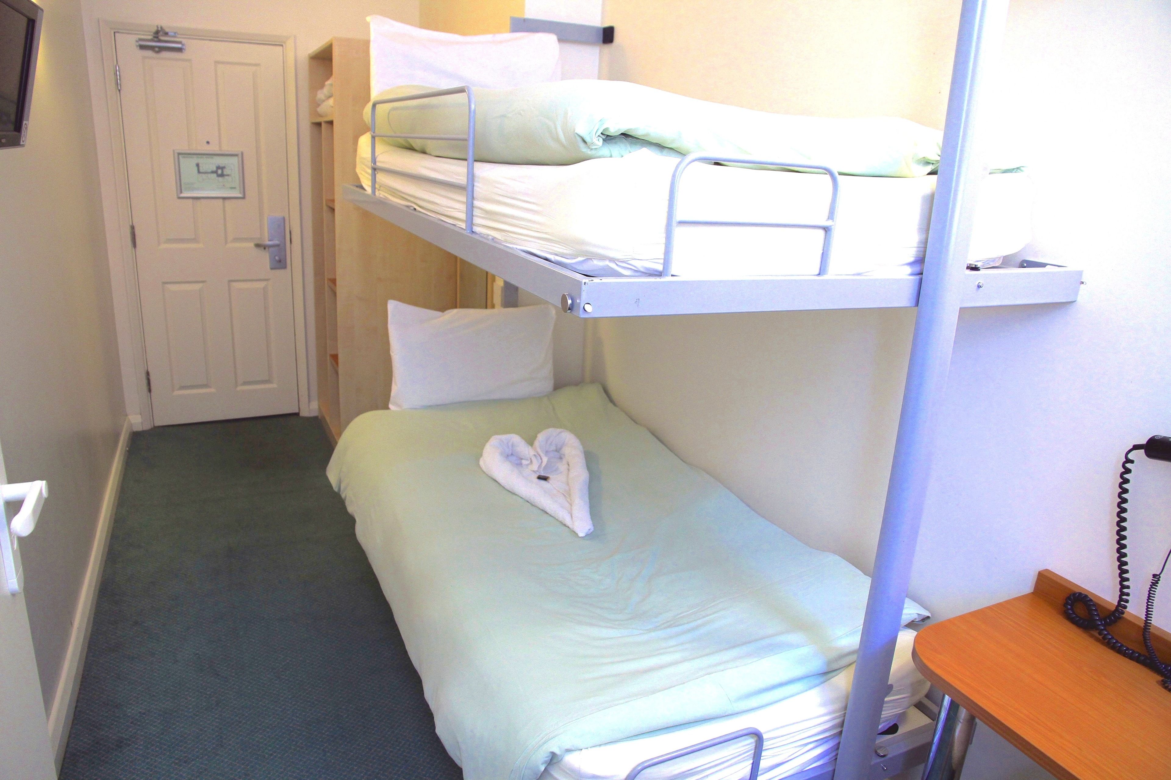 Camera con letto a castello, un asciugamano a forma di cuore sul letto inferiore, una porta con un cartello e un comodino con un telefono.