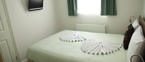 A neatly made bed with a light green bedspread and two decorative white pillows. A flat-screen TV is mounted on the wall above the bed.