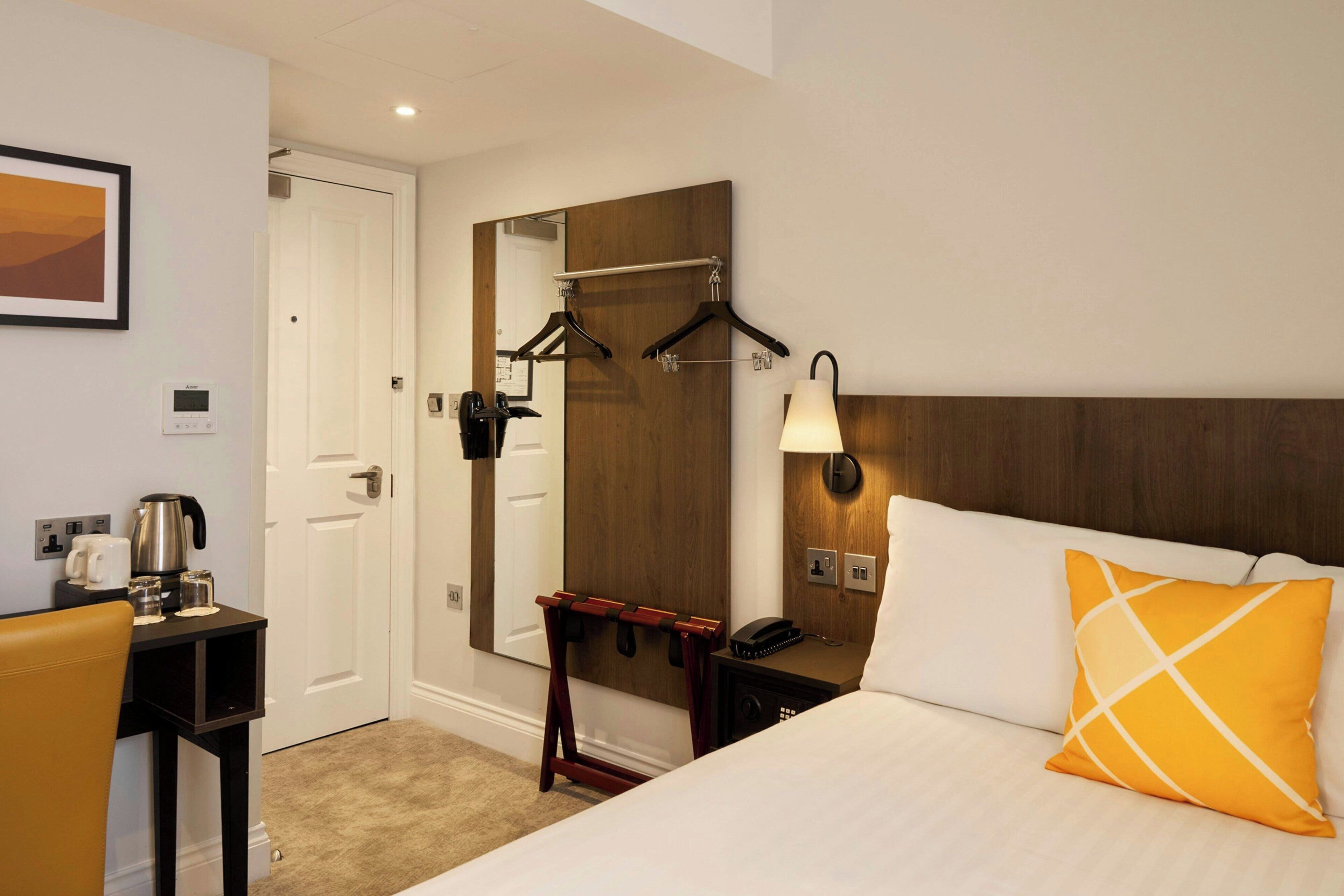 A hotel room with a bed, a desk with a coffee maker, a mirror, and a coat rack.