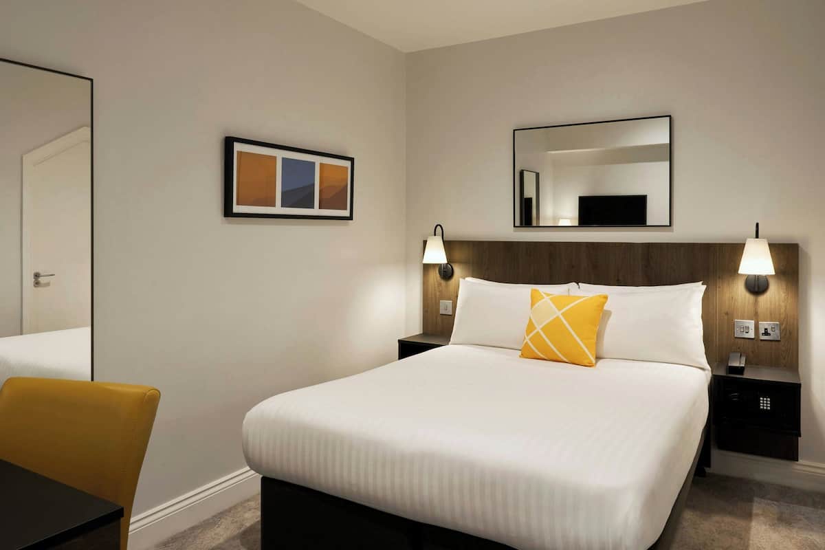 Ein Hotelzimmer mit einem großen Bett, zwei Nachttischen mit Lampen, einem Sessel, einem gerahmten Bild und einem Spiegel an der Wand.