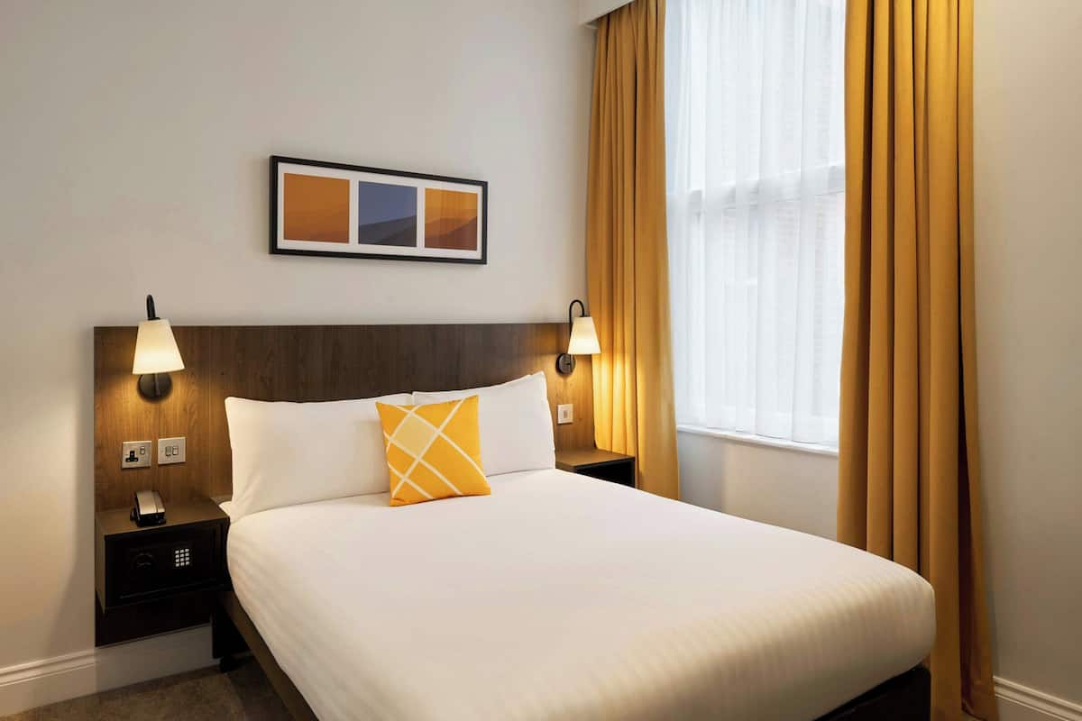 Ein Hotelzimmer mit einem großen Bett, zwei Nachttischlampen, einem gerahmten Bild an der Wand und einem Fenster mit Vorhängen.