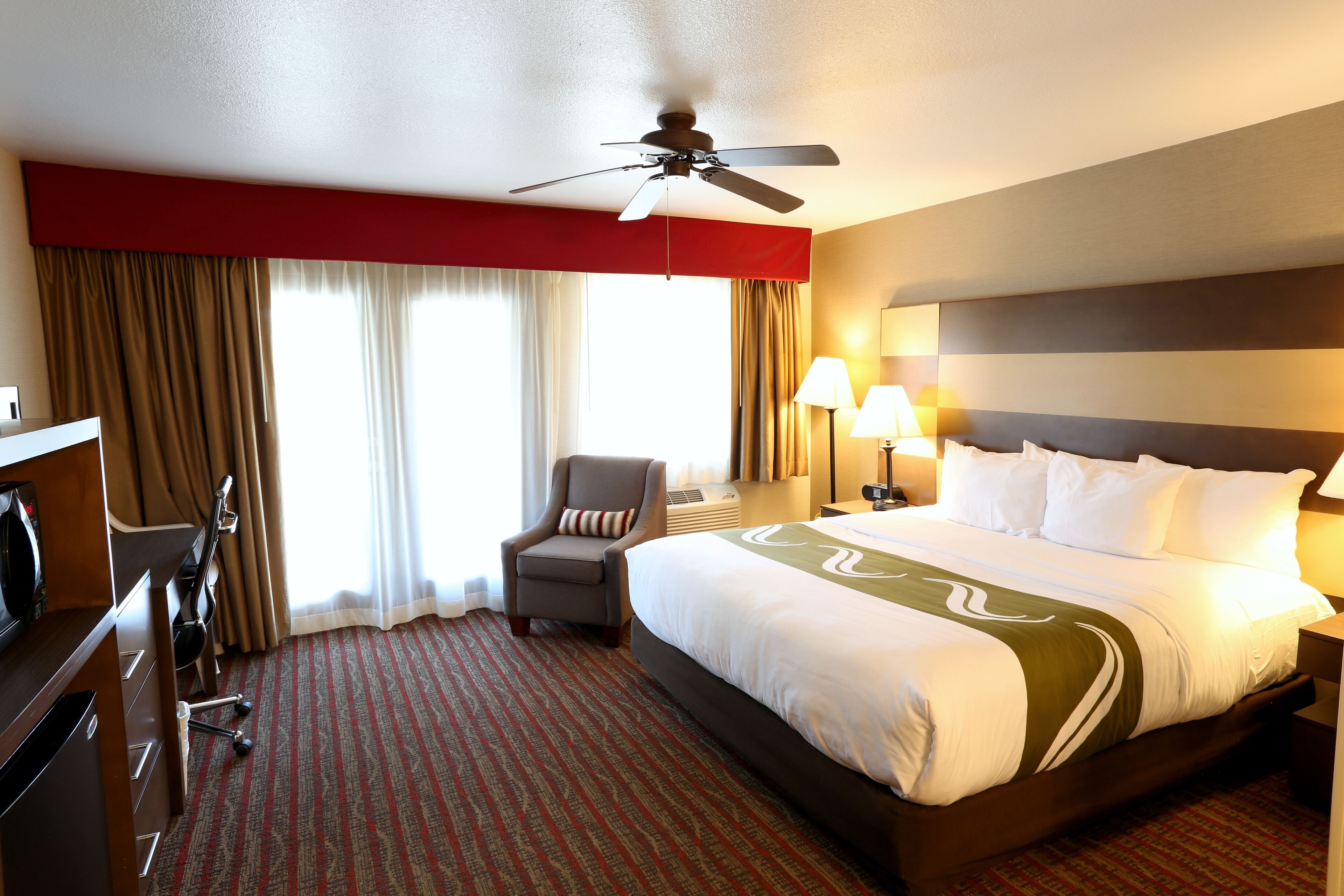 Ein Hotelzimmer mit einem großen Bett, einem Schreibtisch mit Stuhl, einem Deckenventilator und einem Fenster mit Vorhängen.