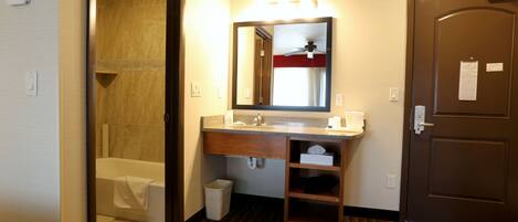 A hotel bathroom with a shower, sink, mirror, and a door.