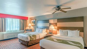 A hotel room with two beds, a ceiling fan, a window with curtains, a wall-mounted heater, and a red carpet.