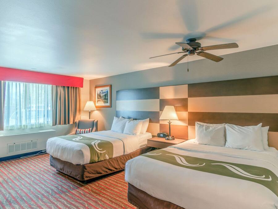 A hotel room with two beds, a ceiling fan, a window with curtains, a wall-mounted heater, and a red carpet.