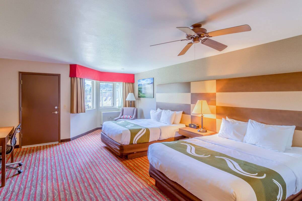 Habitació d'hotel amb dos llits, un escriptori, una cadira, un ventilador de sostre i una finestra amb cortines.