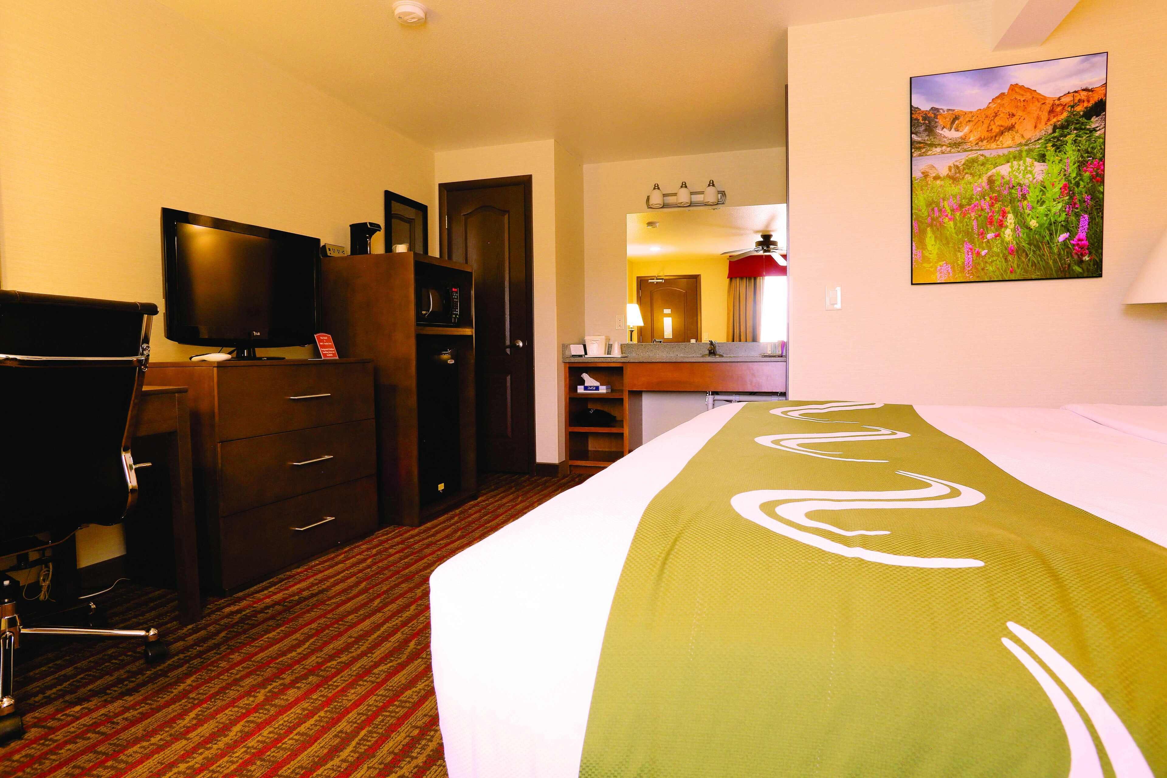 Una camera d'albergo con un letto, una scrivania con un computer, una televisione, un armadio e un bagno visibile attraverso una porta aperta.