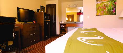 A hotel room with a bed, a desk with a computer, a TV, a wardrobe, and a bathroom visible through an open door.