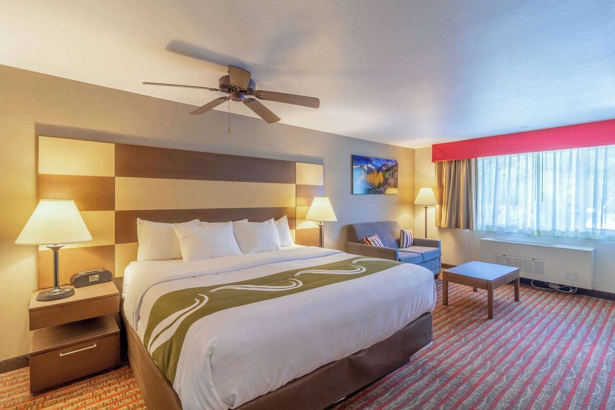 Una habitació d'hotel amb un llit gran, dues taules de nit amb làmpades, un escriptori, una cadira, un ventilador de sostre i una finestra amb cortines.