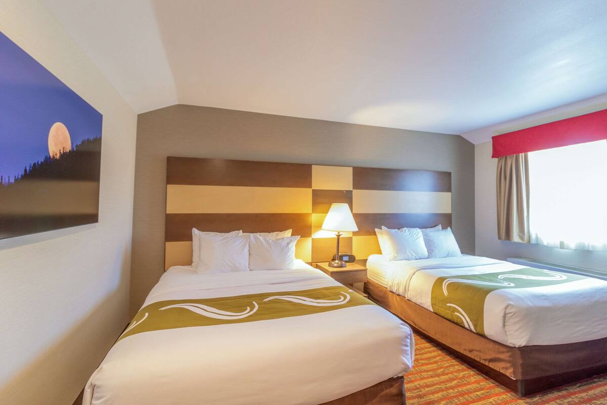 Habitació d'hotel amb dos llits, una taula de nit amb una làmpada, un quadre d'un paisatge a llunars i una finestra amb cortines.