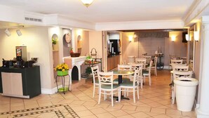 A spacious dining area with multiple tables and chairs, a kitchen area with a coffee machine, and a small seating area with a fireplace. - The Grand by Capital O Texarkana I-30 (Texarkana)