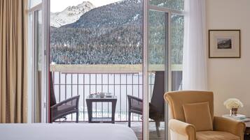 Habitación de hotel con cama, una silla, una mesita con tetera y vistas a montañas nevadas desde un balcón.