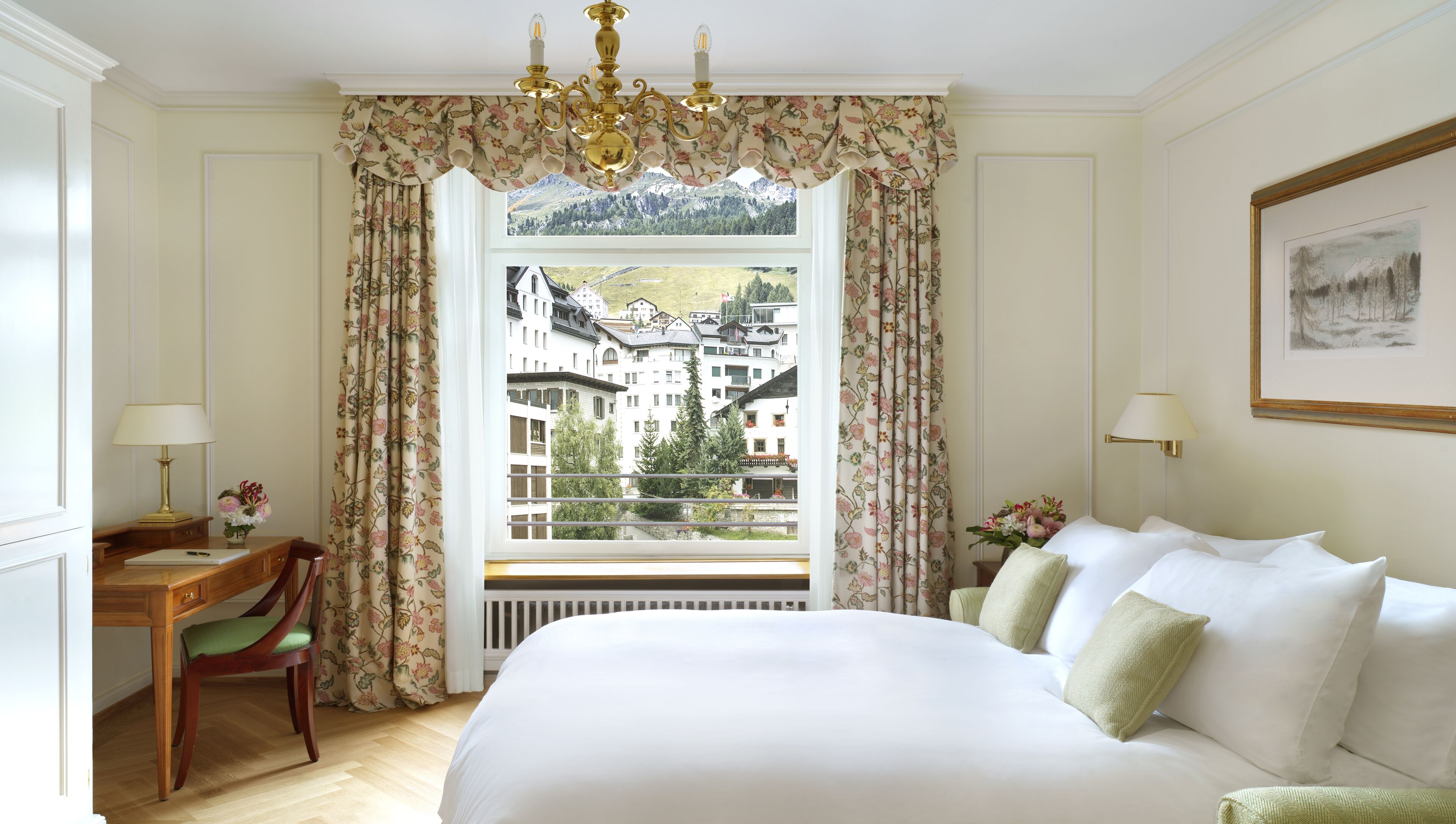 A hotel room with a large bed, a desk with a lamp, a chair, and a view of a mountainous landscape through a window with floral curtains.