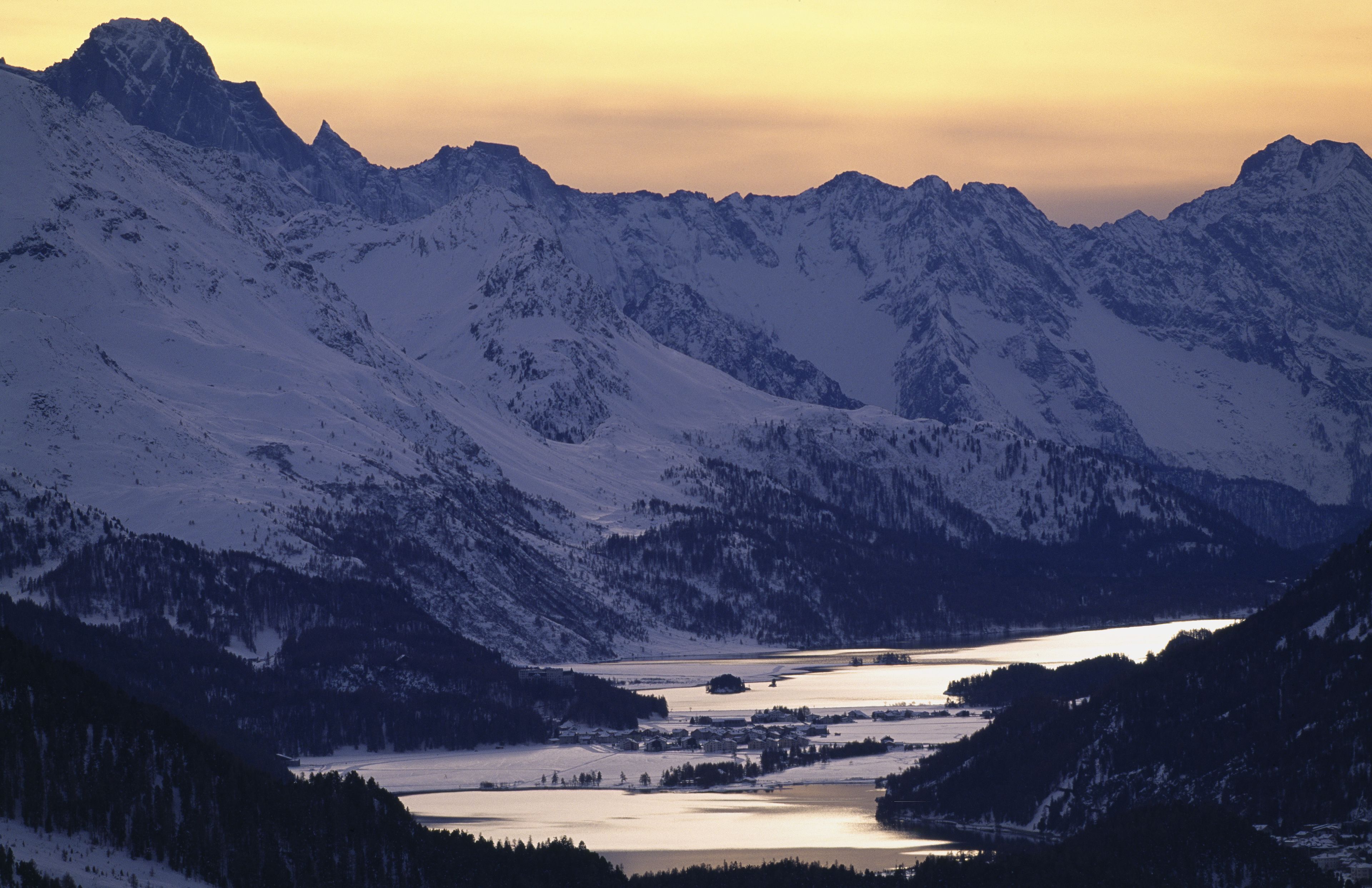 Eine Berglandschaft mit schneebedeckten Gipfeln und einem Tal mit einem Gewässer.