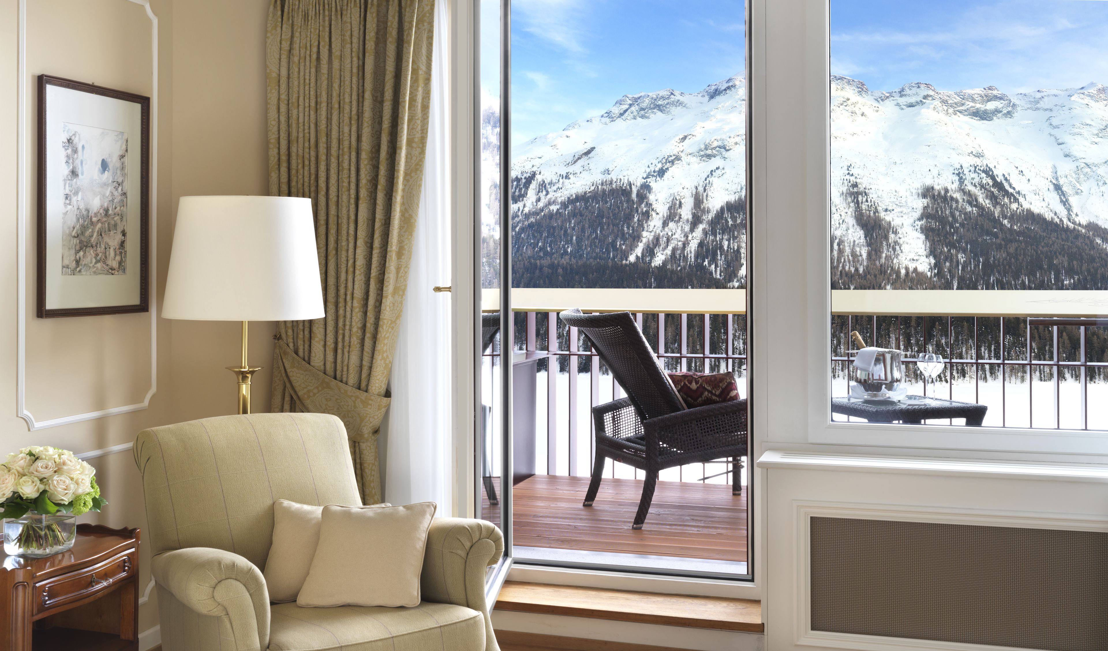 A room with a view of snow-covered mountains, a chair, a lamp, and a vase of flowers.