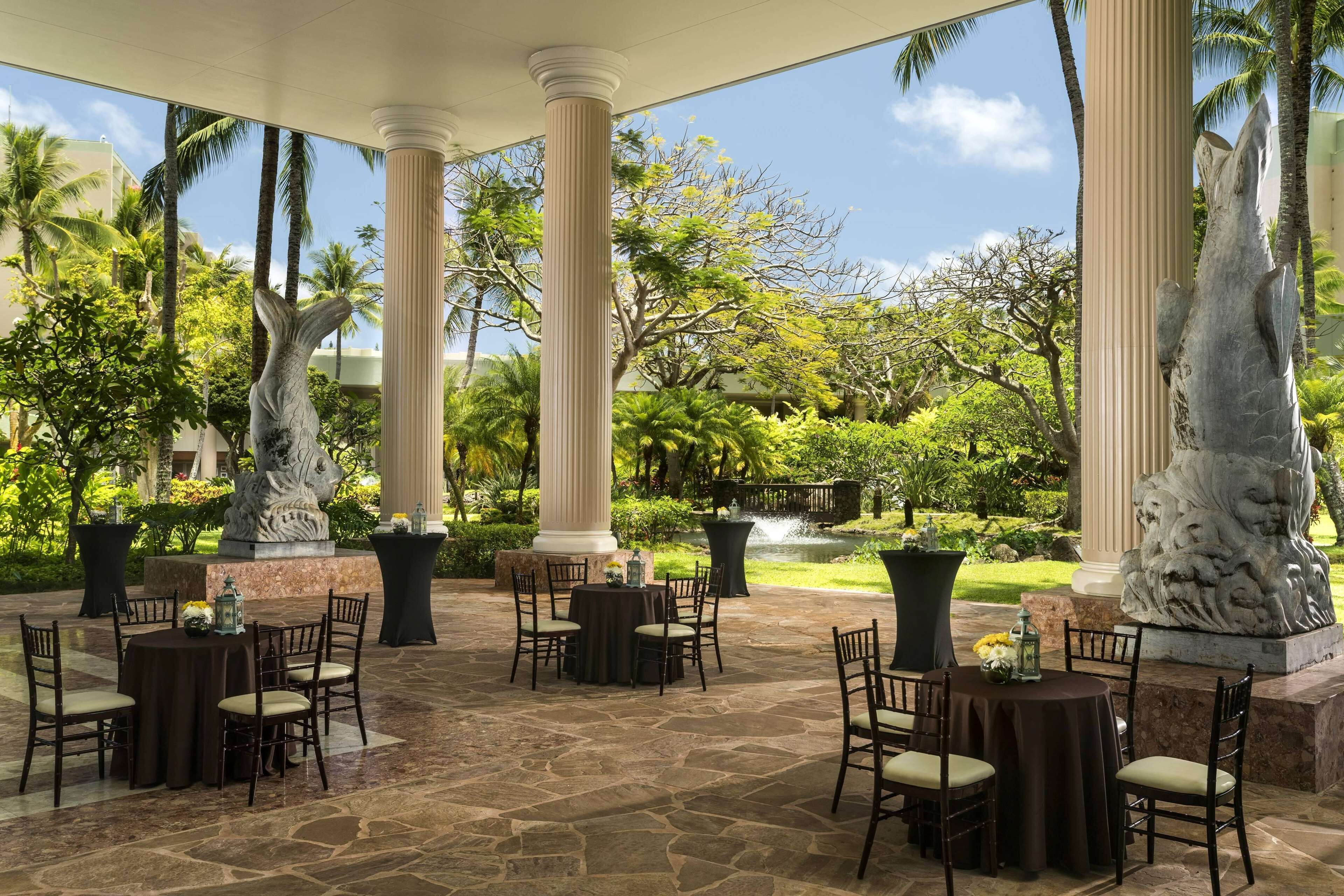 Une terrasse extérieure avec un sol en pierre, des tables et des chaises rondes, entourée de palmiers et de colonnes.