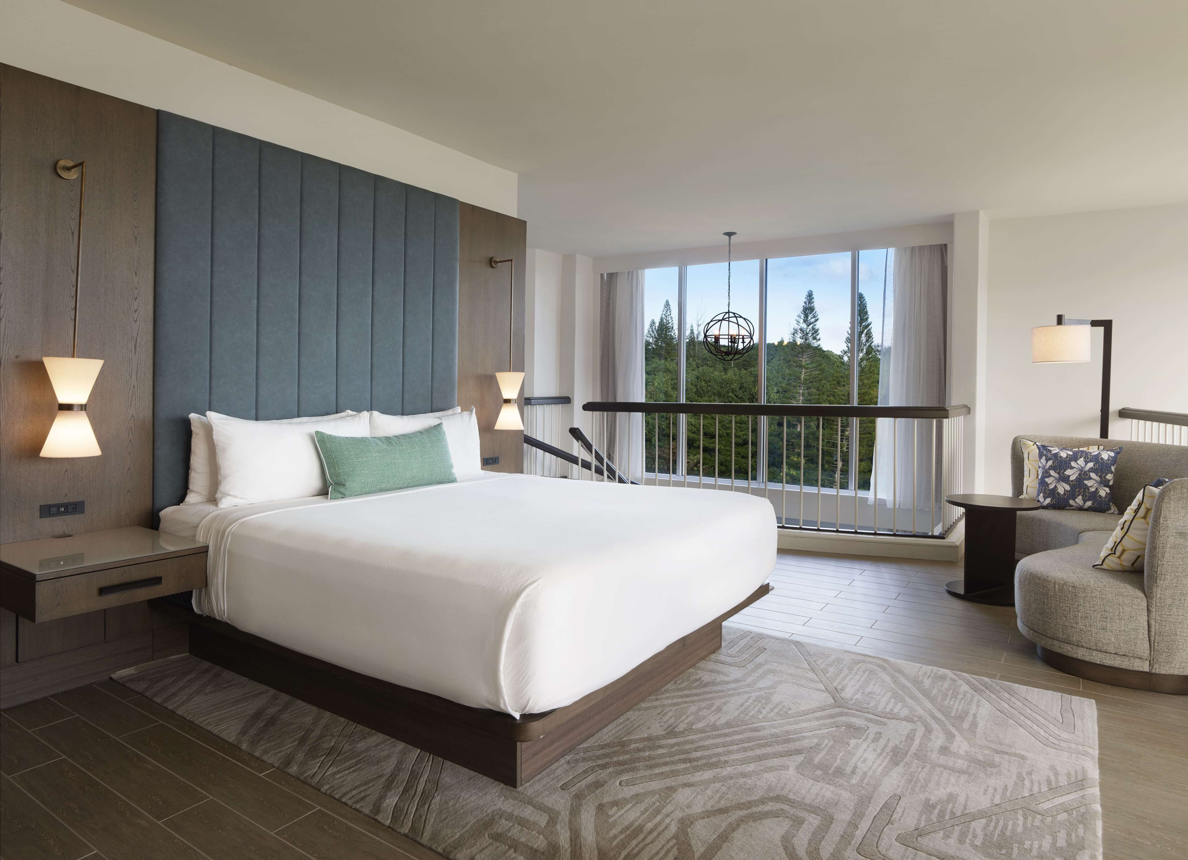 Ein modernes Hotelzimmer mit einem großen Bett, einem Balkon mit Geländer und Blick auf Bäume.