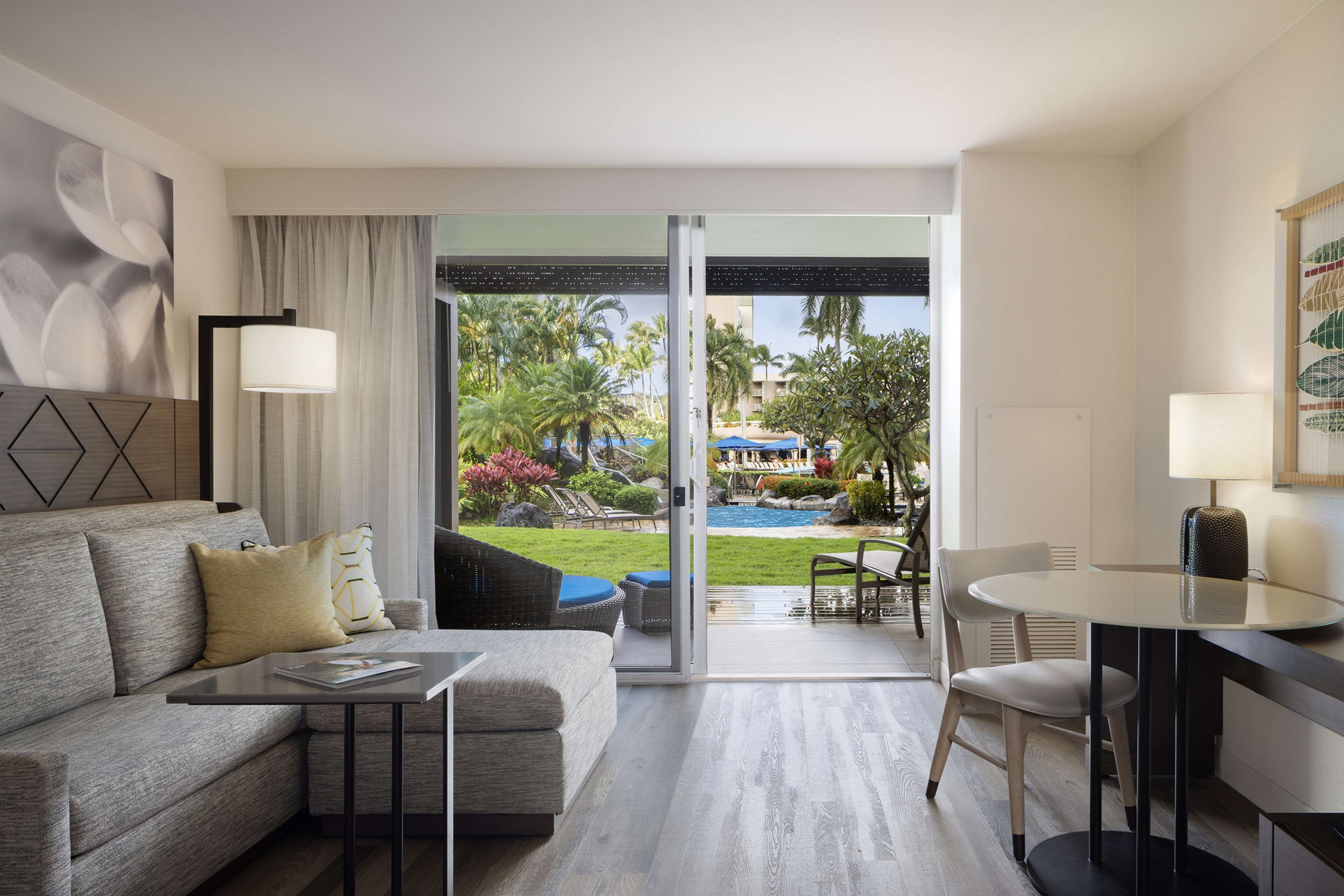 Una habitación de hotel moderna con sofá, una mesita, un área de comedor y un ventanal corredero que da a una piscina rodeada de palmeras y vistas al complejo turístico.