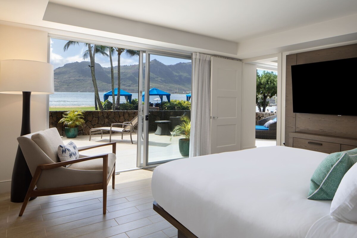 A hotel room with a large bed, a TV, a chair, a lamp, and a view of the beach and mountains.