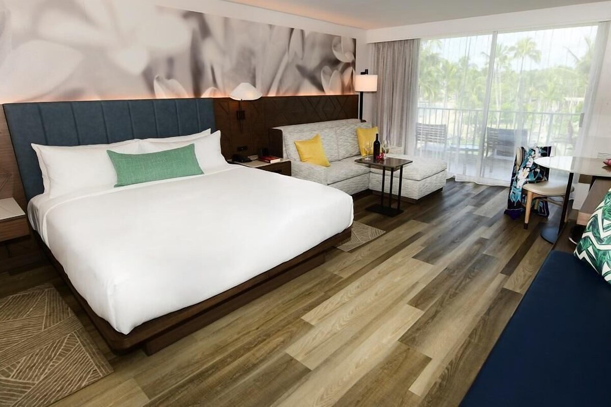 A hotel room with a large bed, a sofa, a small table, and a balcony with a view of palm trees.