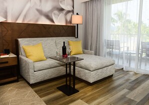 A modern hotel room with a grey sofa, two yellow cushions, a small table with a bottle and glasses, a wooden headboard, and a view of palm trees through sheer curtains.