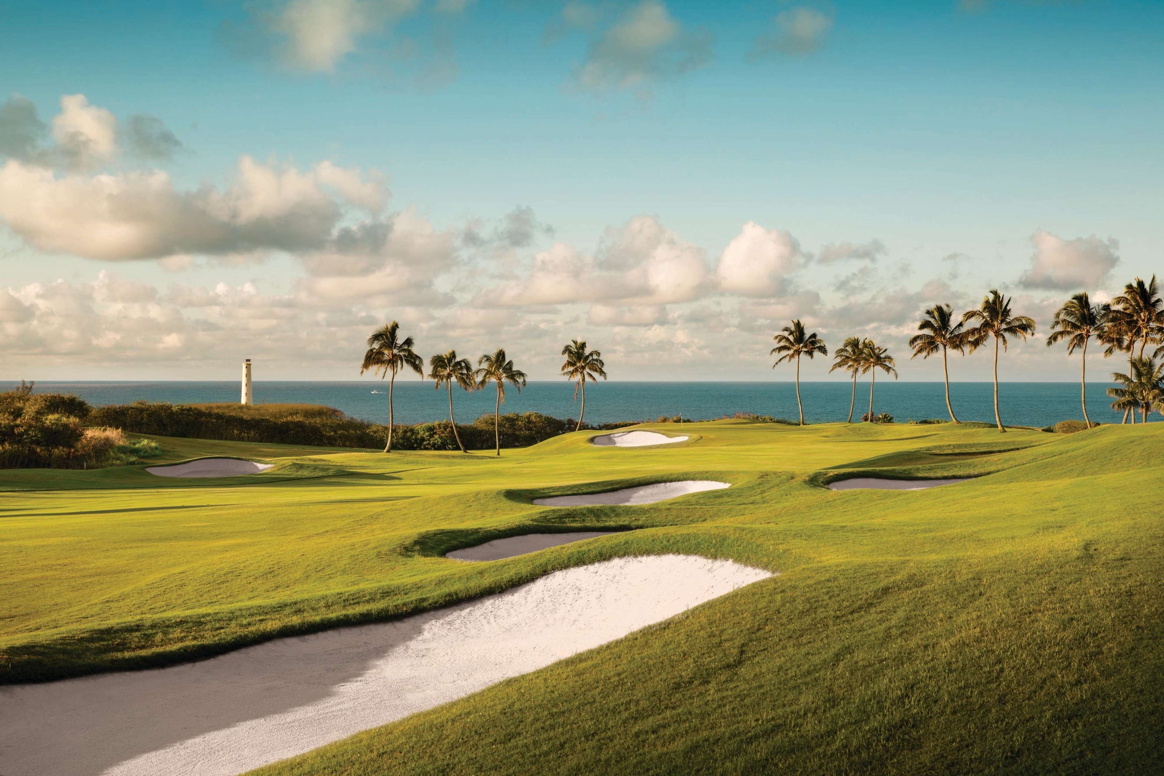 Un campo de golf con búnkeres de arena, palmeras y un faro al fondo.