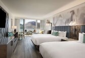 A hotel room with two beds, a desk, a chair, and a large window with a view of the mountains.