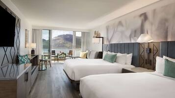 Habitación de hotel con dos camas, un escritorio, una silla y un amplio ventanal con vistas a las montañas.
