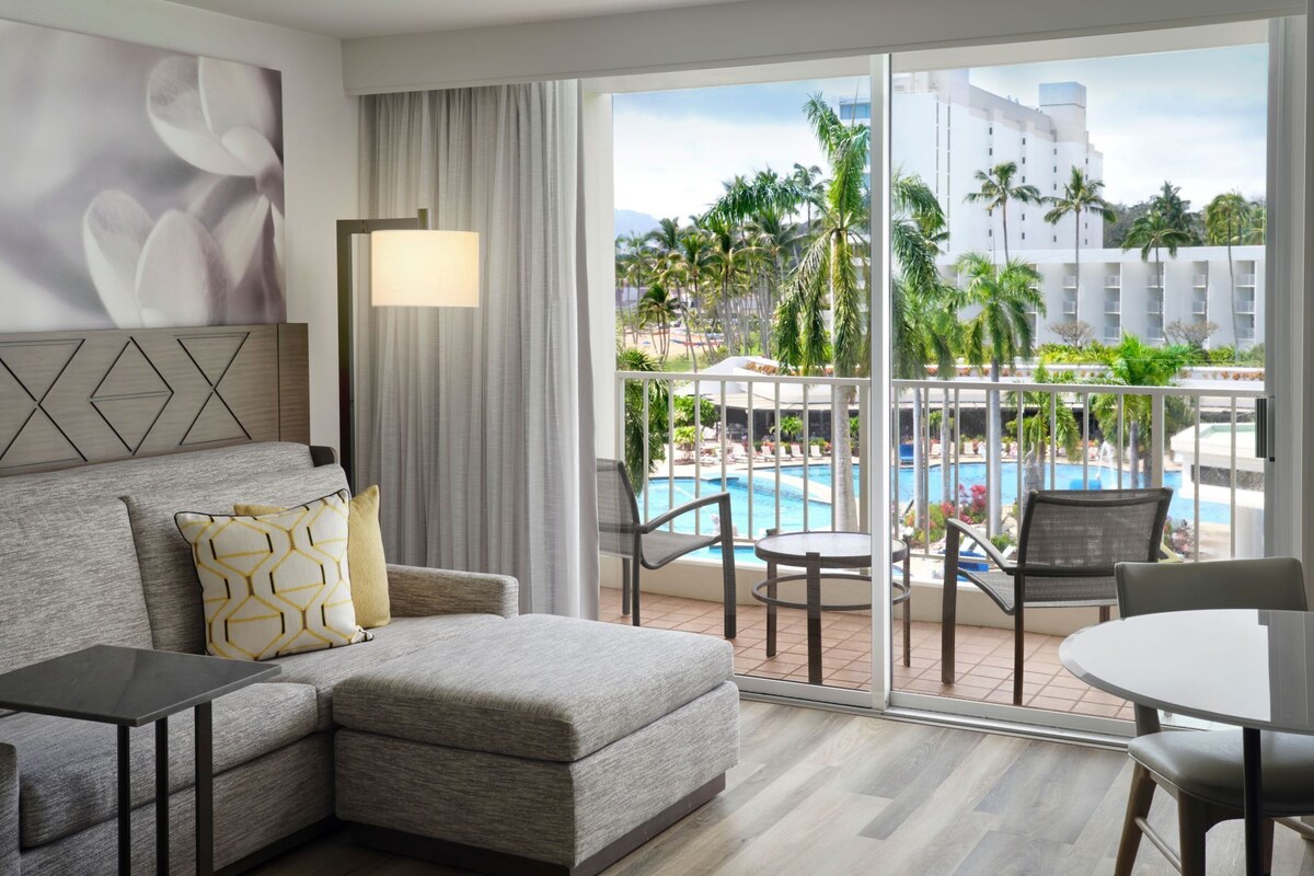 A hotel room with a sofa, a table, a view of a pool and palm trees, and a large window.