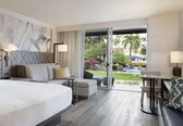 A modern hotel room with a large bed, a sofa, a dining table, and a view of a pool and garden.