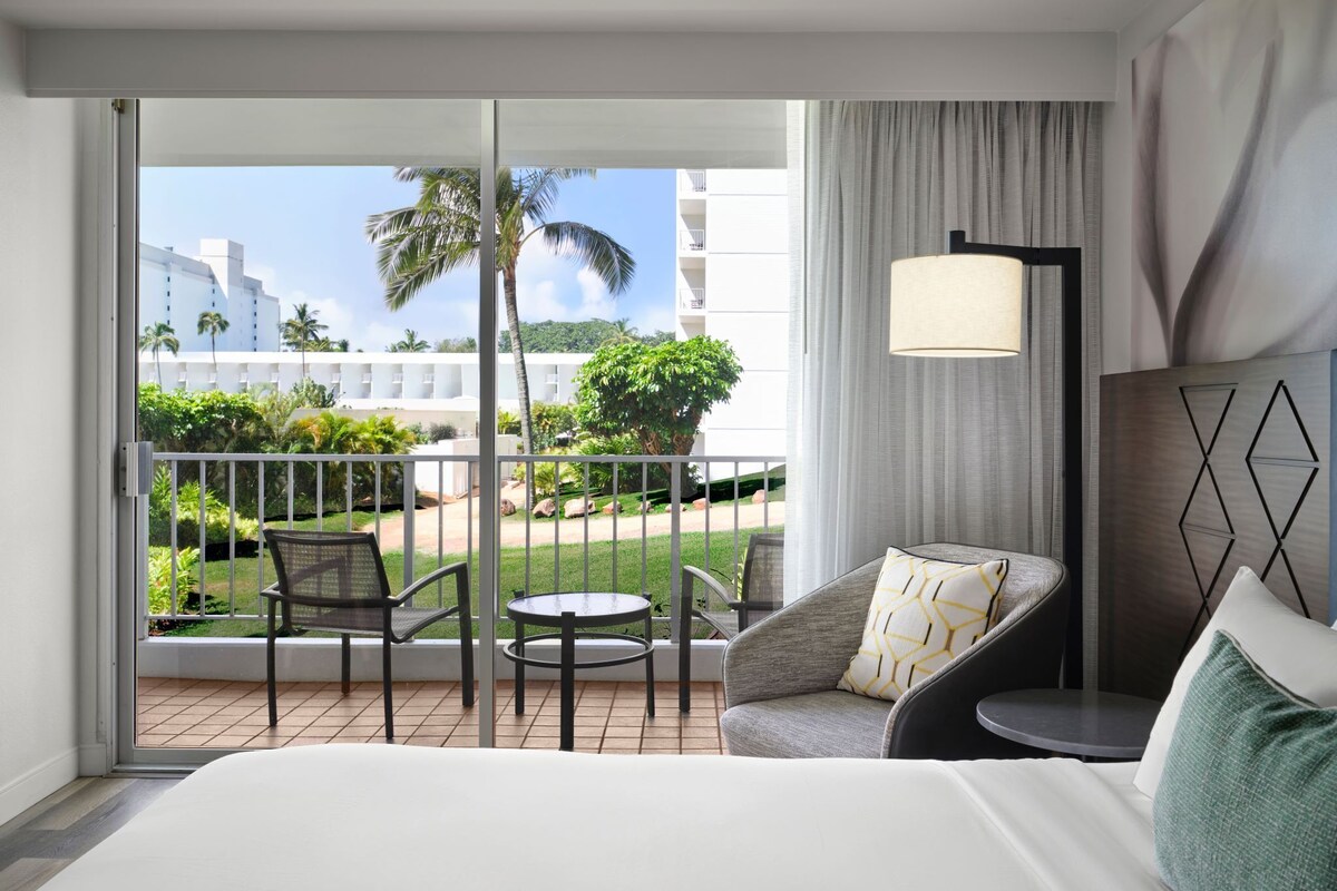 A hotel room with a balcony, a bed, a chair, a table, and a lamp.