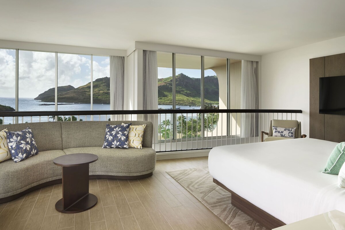 A hotel room with a large bed, a curved sofa, a small table, and a view of the ocean.