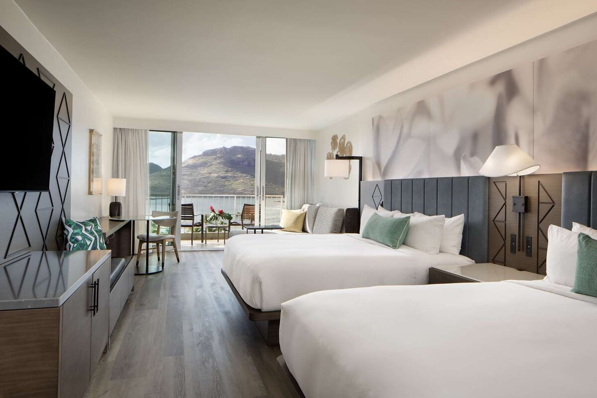 A hotel room with two beds, a desk, a TV, and a balcony with a view of mountains.