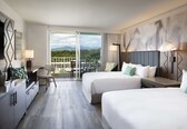 A hotel room with two beds, a desk, a chair, and a balcony with a view of mountains.