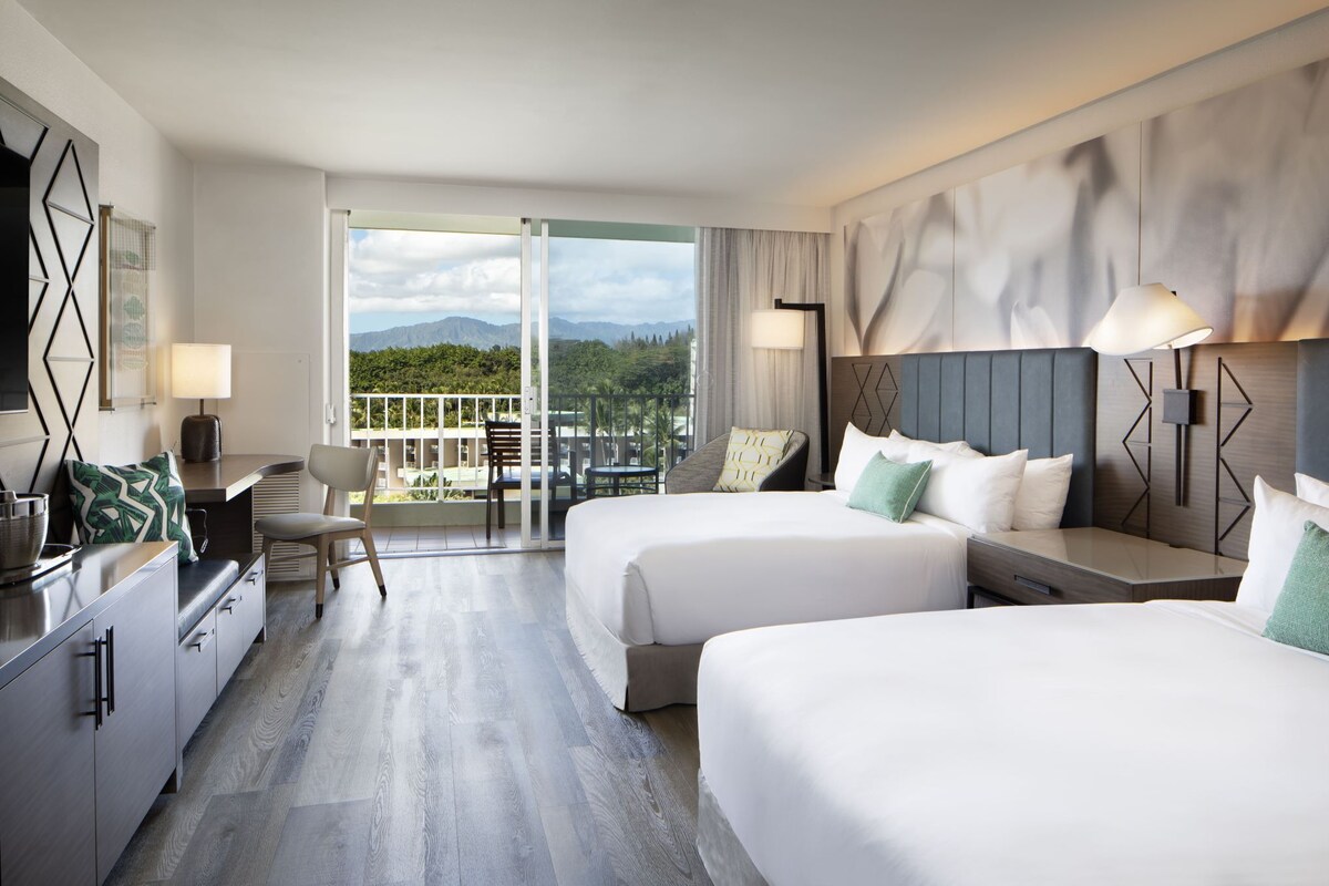 A hotel room with two beds, a balcony, a desk, and a view of the mountains.