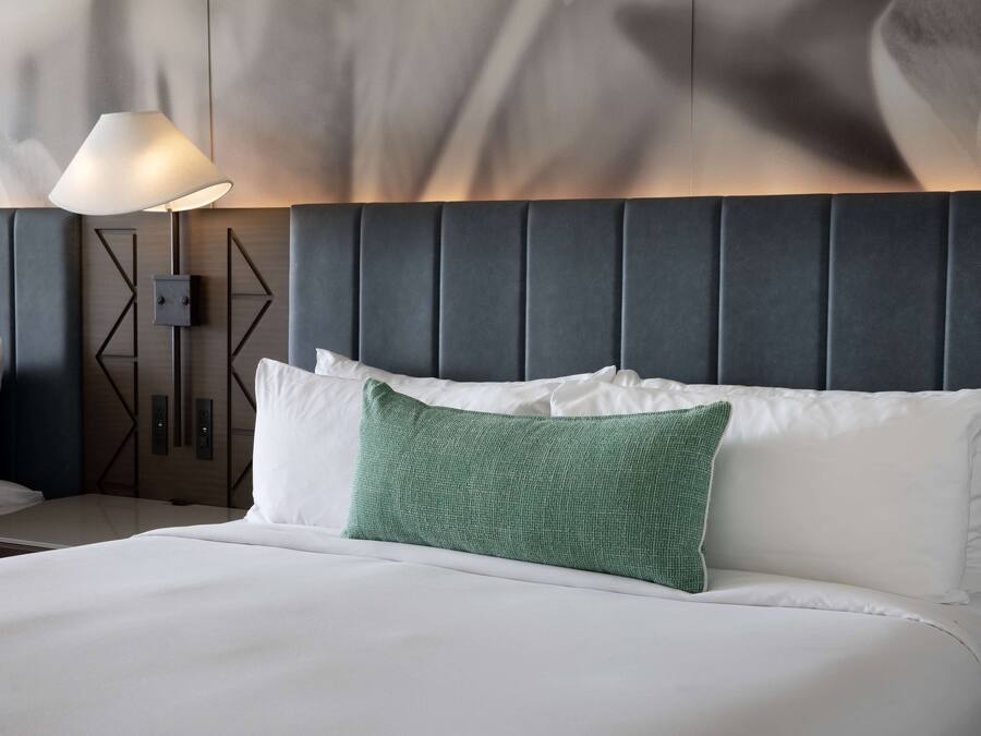 A neatly made bed with white linens and a green accent pillow, a modern lamp, and a geometric wall panel.