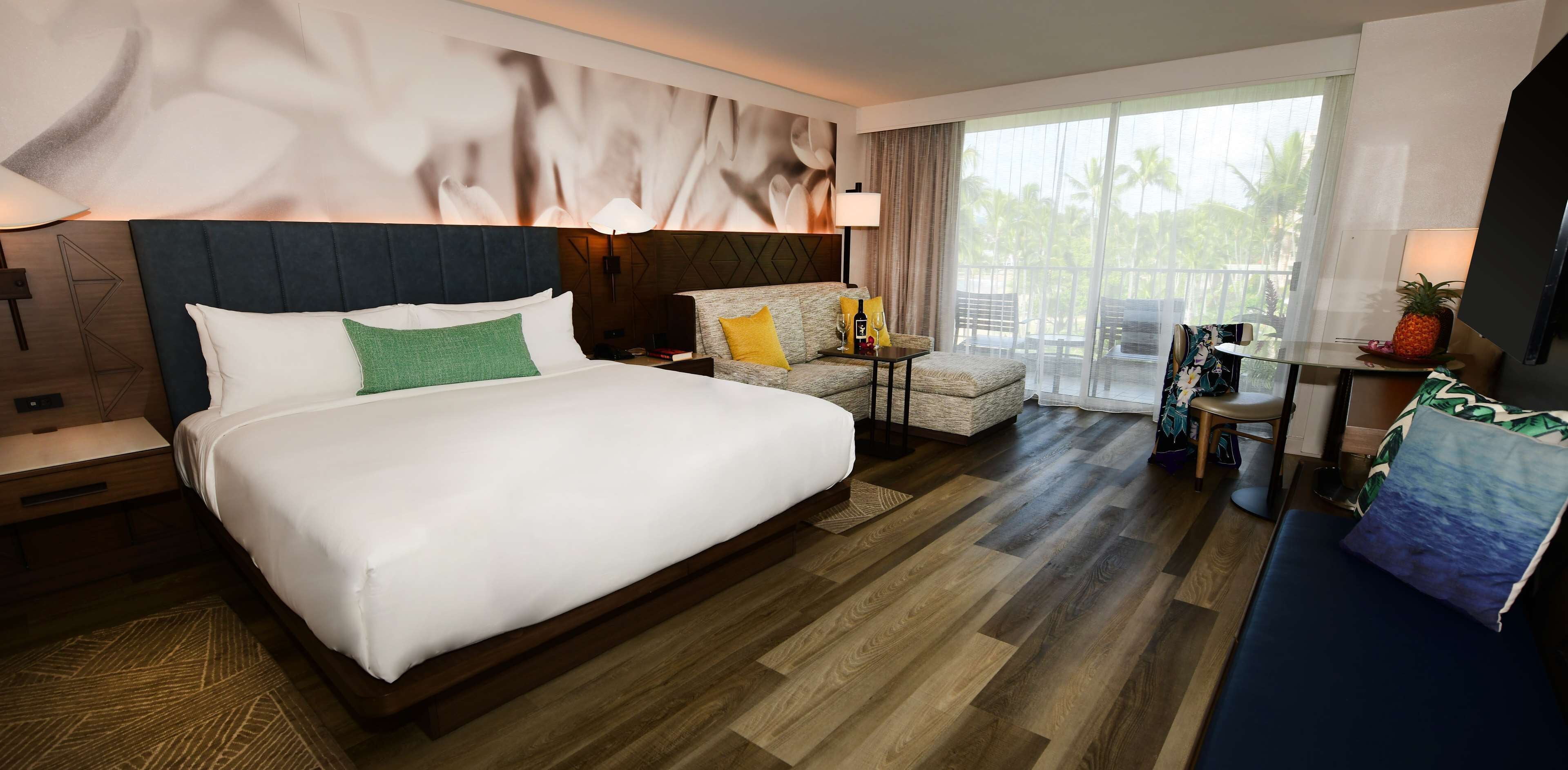 Ein Hotelzimmer mit einem großen Bett, einer Couch, einem kleinen Tisch und einem Balkon mit Blick auf Palmen.