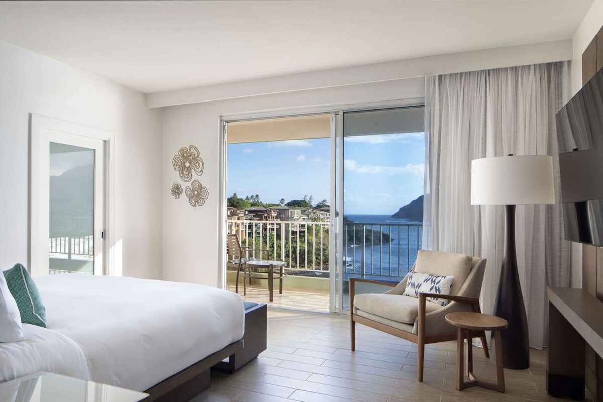 A hotel room with a bed, a chair, a small table, a lamp, and a balcony with a view of the sea.
