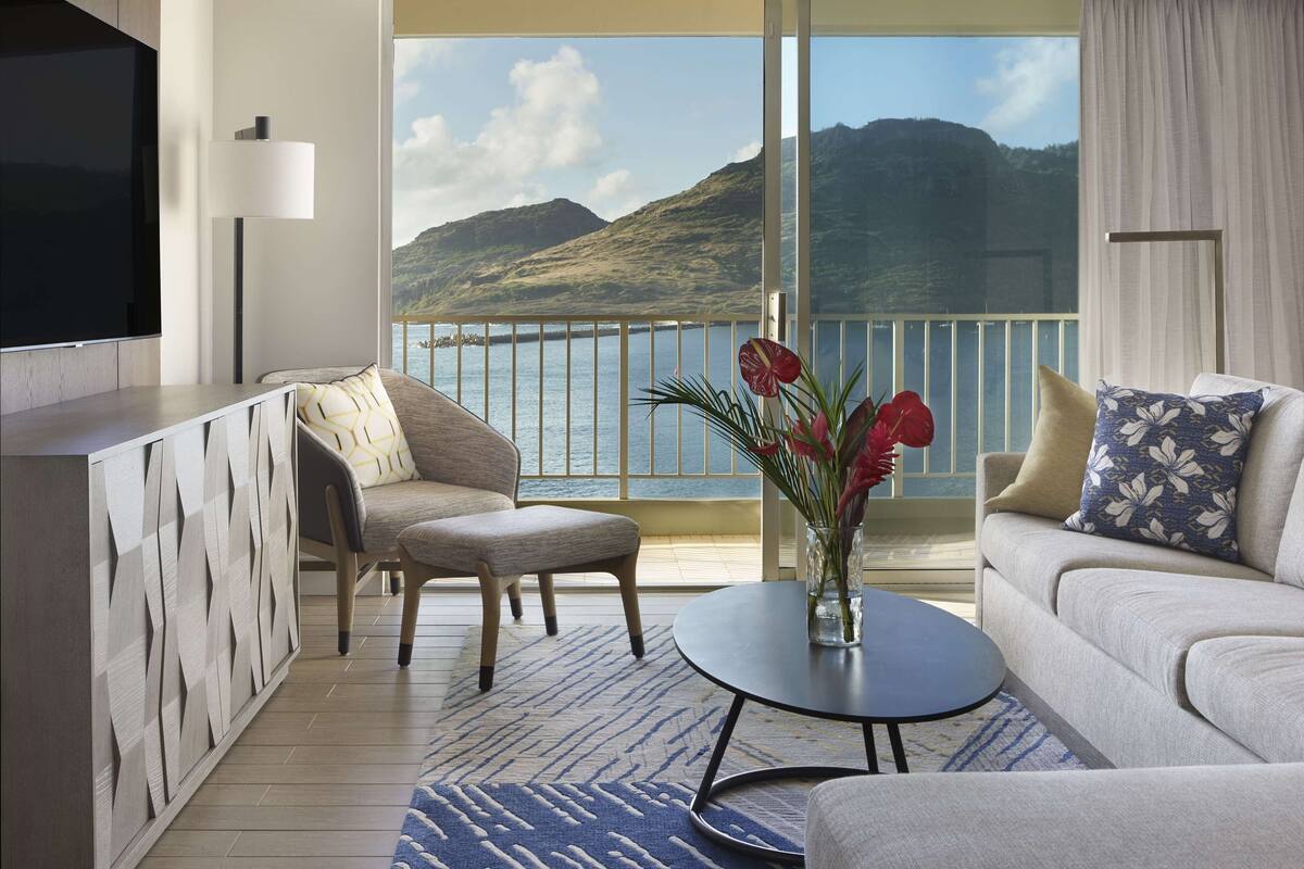 A modern hotel room with a balcony, a flat-screen TV, a sofa, a coffee table, and a decorative vase with flowers.