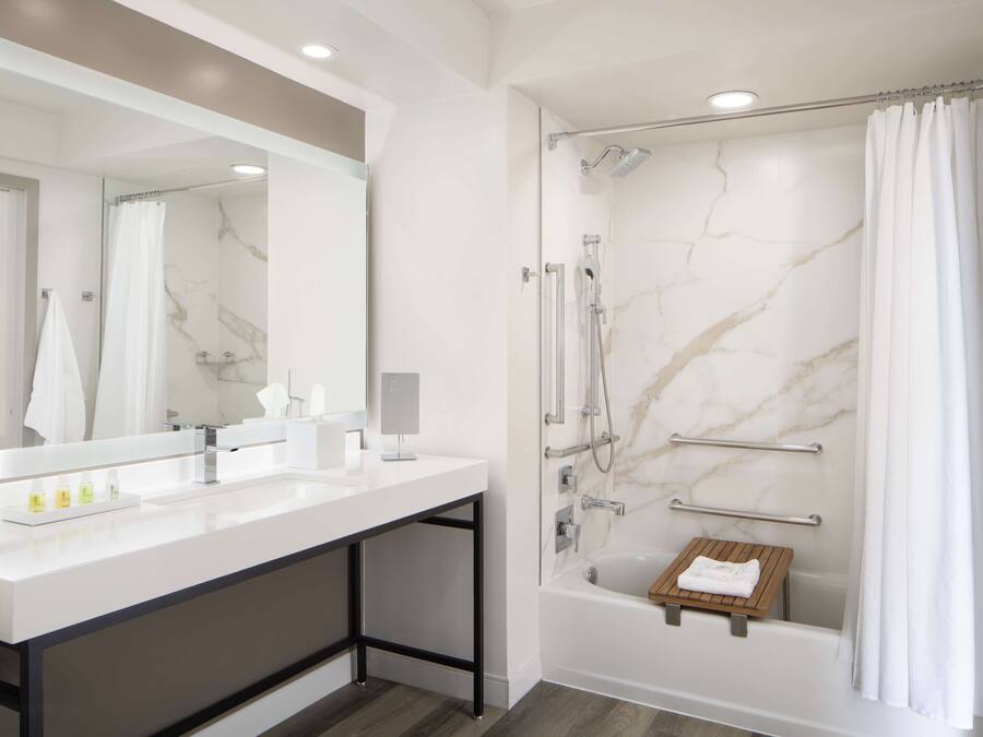 A modern bathroom with a large mirror, two sinks, a shower with a handheld showerhead, and a wooden shelf.