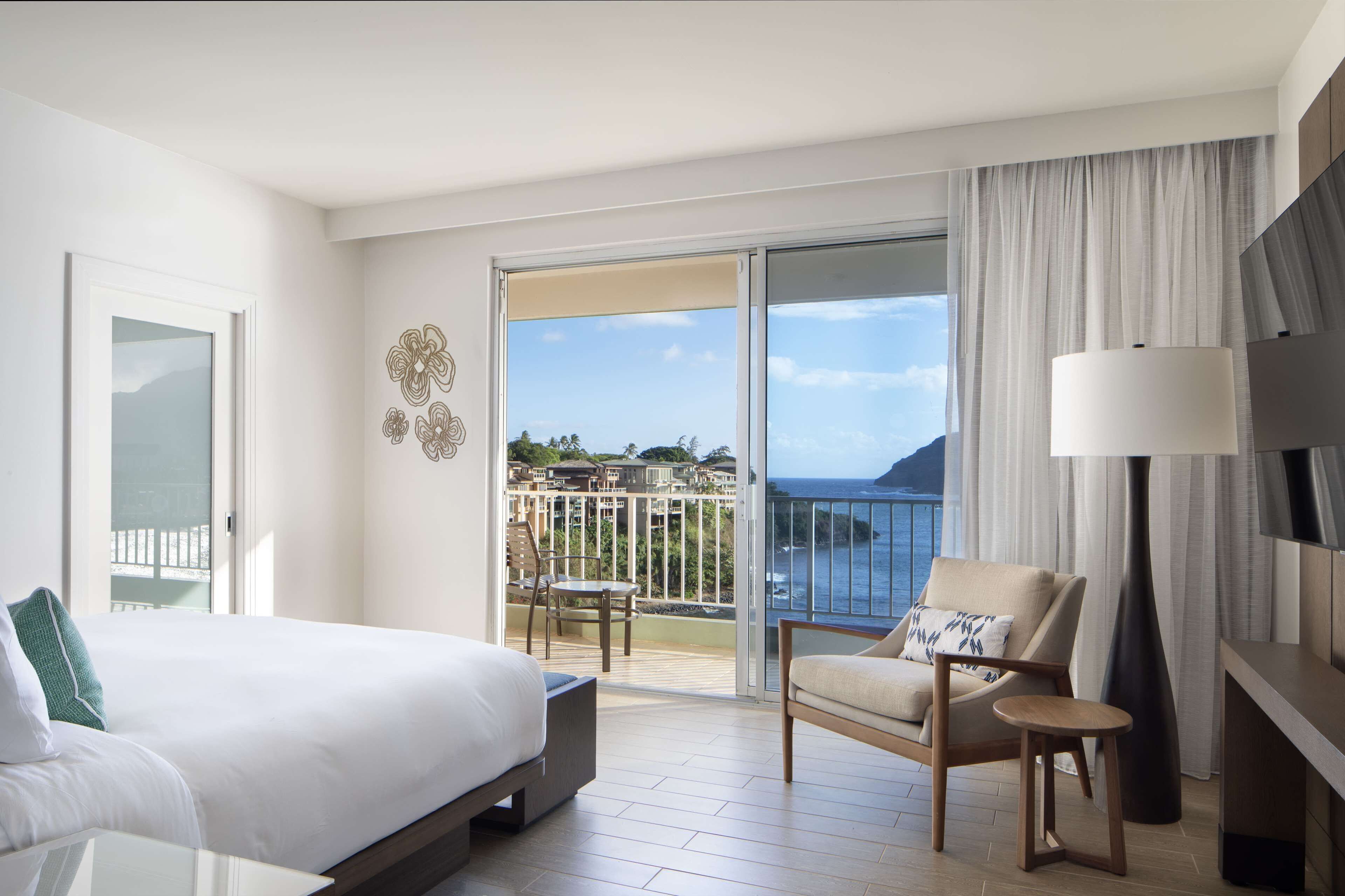 A hotel room with a bed, a chair, a small table, a lamp, and a balcony with a view of the sea.