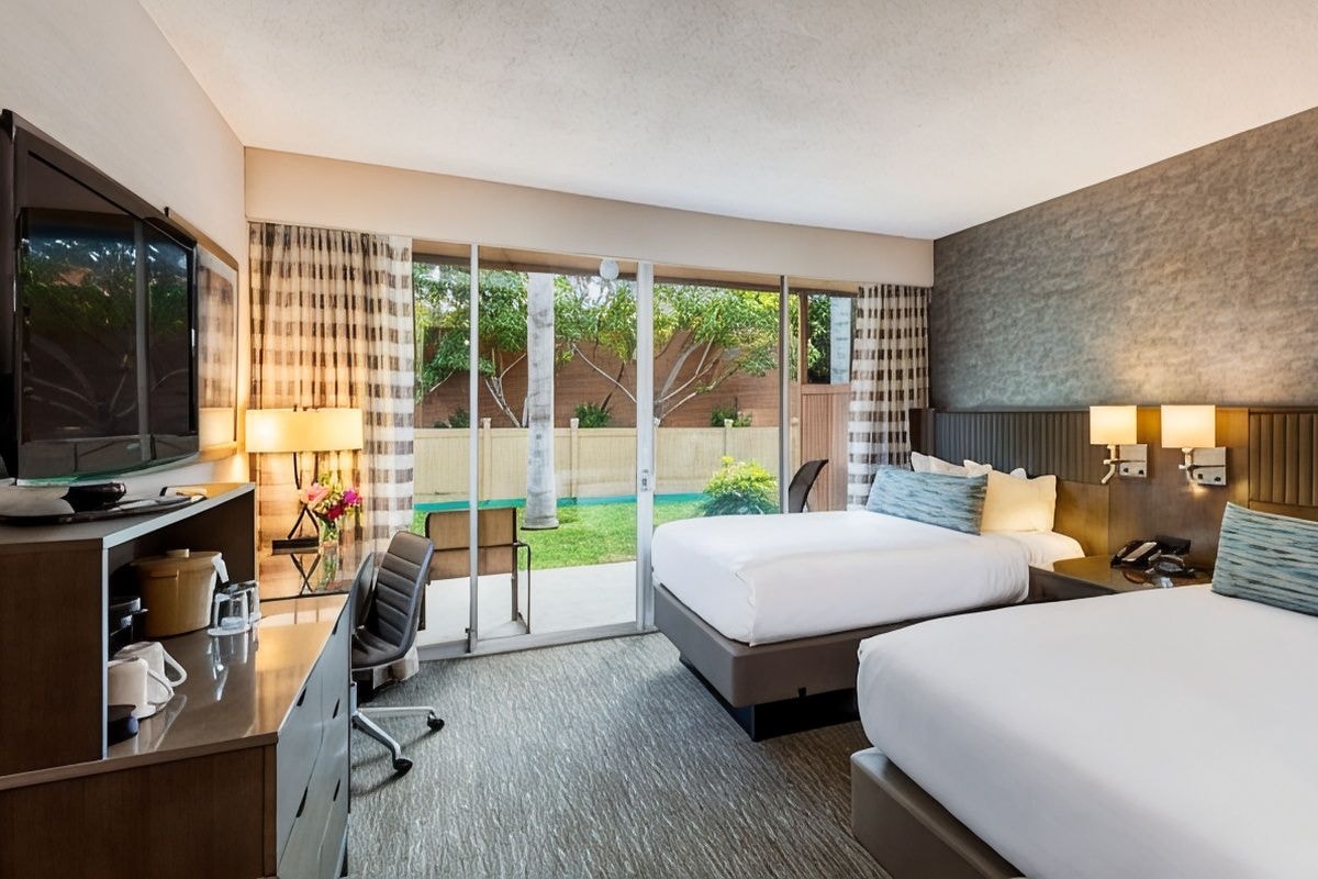 A hotel room with two beds, a desk with a computer, a TV, and a sliding glass door leading to a garden.