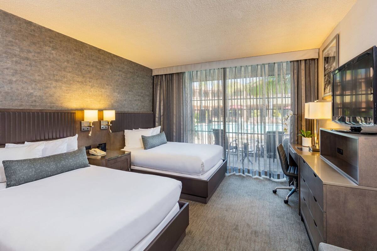 A hotel room with two beds, a desk, a TV, and a view of the outdoors.