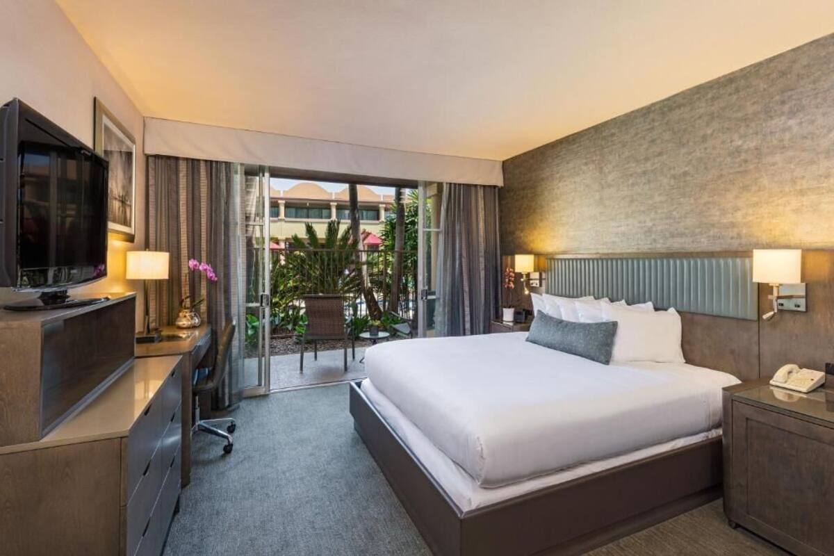 A hotel room with a large bed, a desk with a TV, a phone, and a view of the outdoor area.