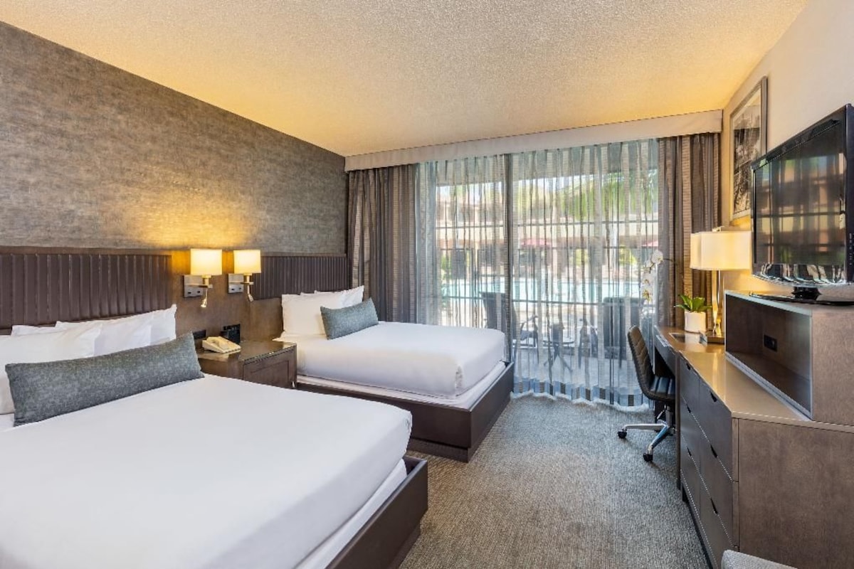 A hotel room with two beds, a desk, a TV, and a large window with curtains.